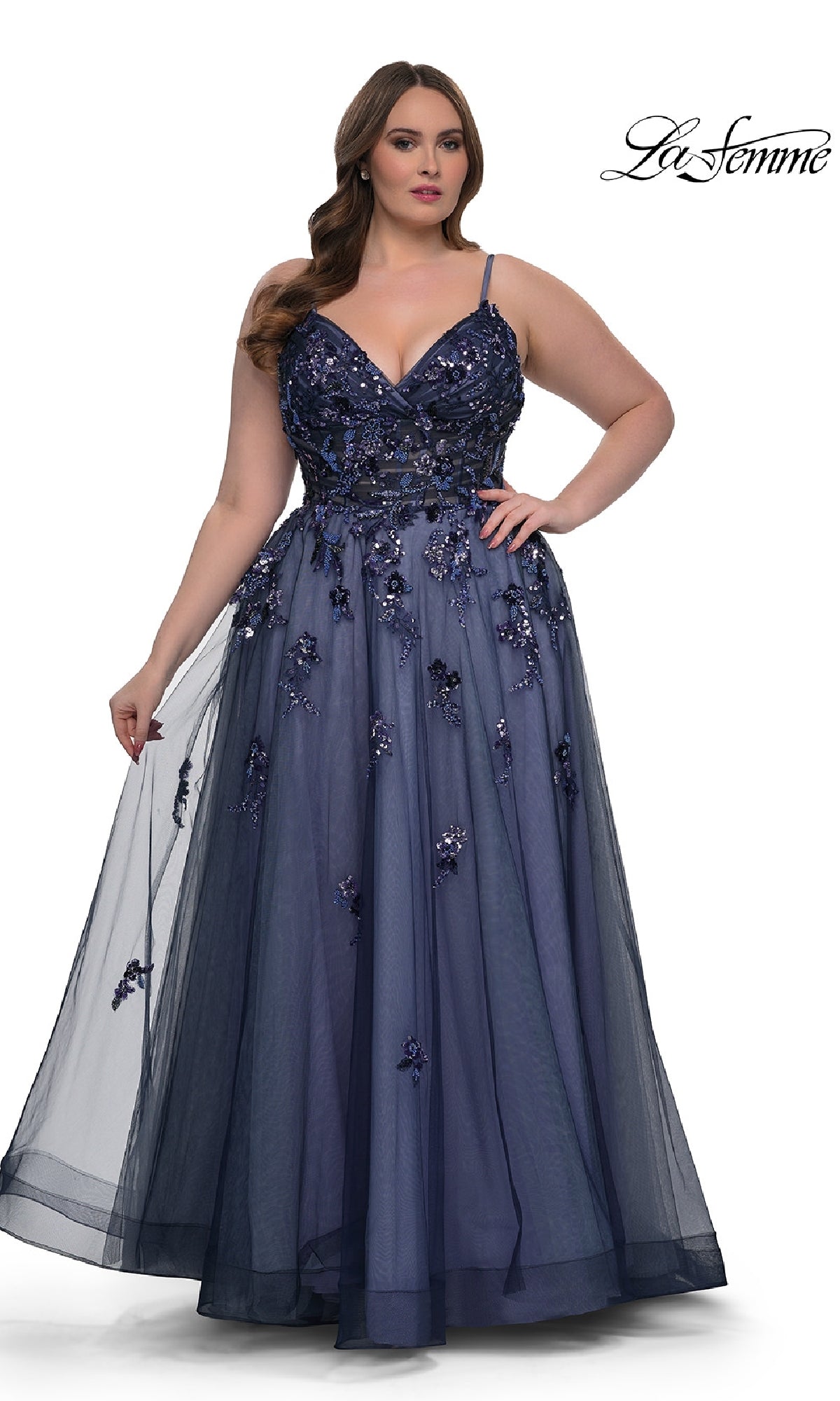 A woman wearing the Navy Plus-Size Prom Dress: La Femme 33061, featuring a floor-length A-line skirt, sequin beaded floral applique, and spaghetti straps, stands while holding out part of the elegant skirt.